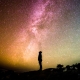 Die Silhouette eines zum Himmel emposchauenden Menschen. Er schaut in den Nachthimmel, der tausende Sternen zeigt und die Milchstraße